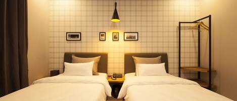 Habitación de hotel con dos camas, una mesita de noche con lámpara, perchero y pared decorada con cuadros.