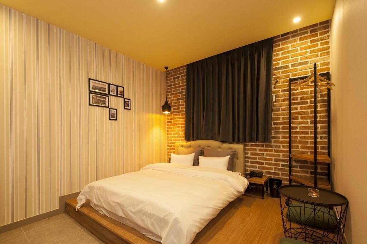 A bedroom with a bed, a nightstand, a chair, a small table, a lamp, and a wall with a striped pattern and a brick wall section.