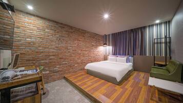 A hotel room with a brick wall, a bed with white linens, a desk with a chair, and a green armchair.