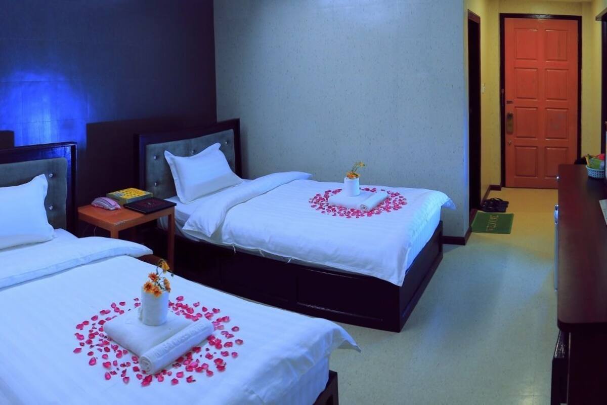 Quarto de hotel com duas camas, cada uma com roupa de cama branca e um arranjo floral decorativo. Tem uma mesa de cabeceira com uma luminária e uma pequena mesa com um livro. O quarto tem uma parede escura e uma porta de madeira.