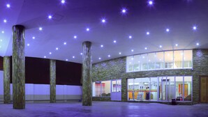 Un vestíbulo de hotel con diseño moderno, que presenta una fachada de vidrio curvo, una zona de recepción y un interior espacioso con techo alto y varias lámparas.