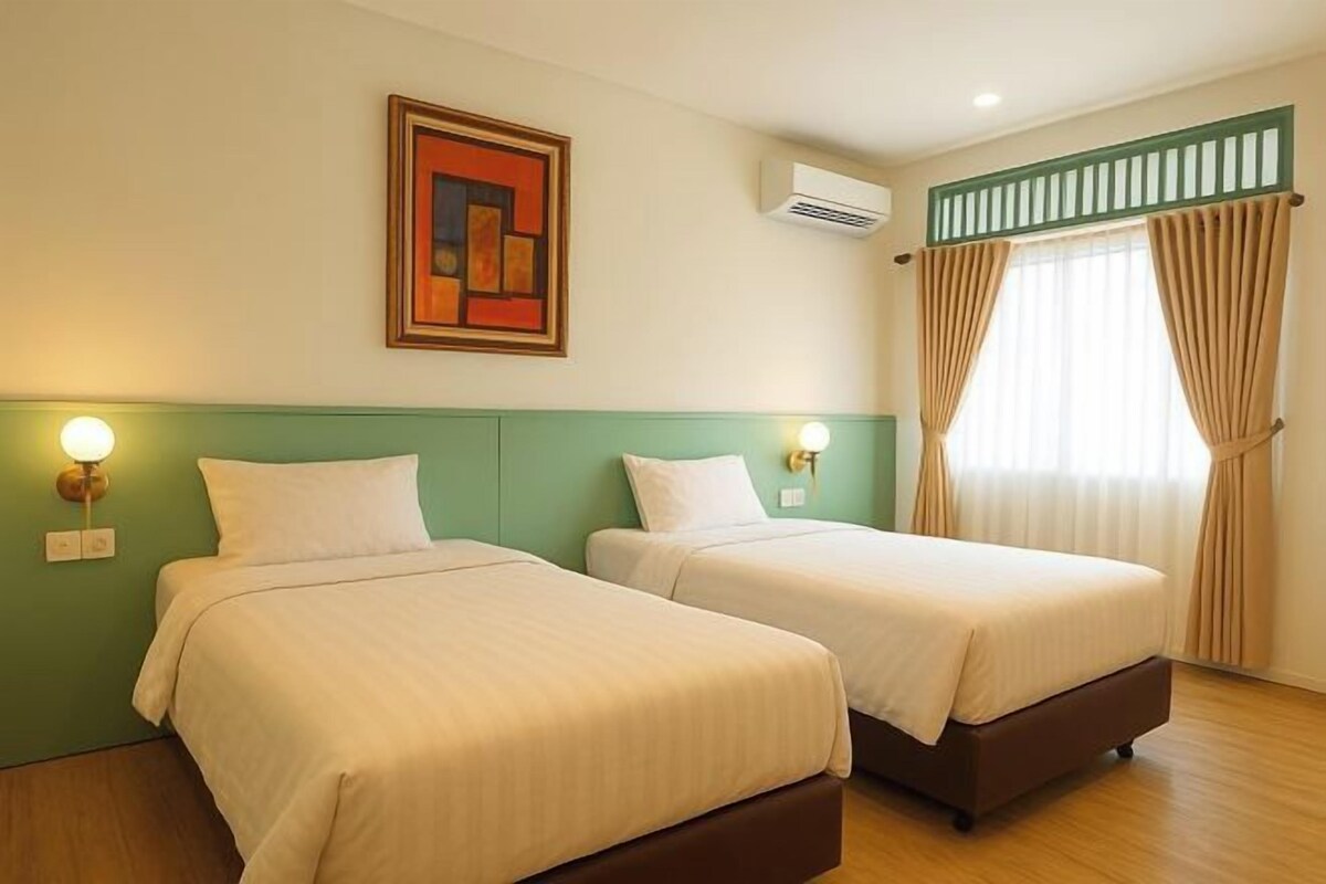 Habitación de hotel con dos camas, cabecera verde, un cuadro en la pared y una ventana con cortinas.