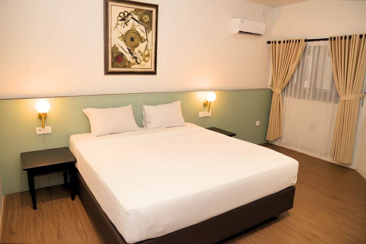 A neatly made bed with white linens, two bedside tables with lamps, a wall-mounted air conditioner, and a window with beige curtains.