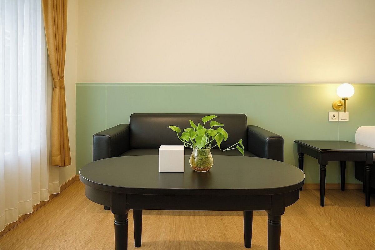 A room with a round black table, a white vase with green plants, a black sofa, and a small table with a lamp.