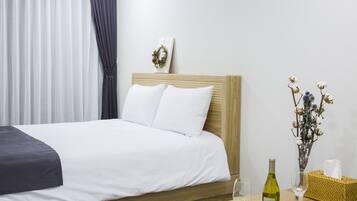 Un lit bien fait, avec du linge de lit blanc, une tête de lit en bois, un vase contenant des fleurs séchées, une bouteille de vin et un verre posés sur une table de chevet en bois.