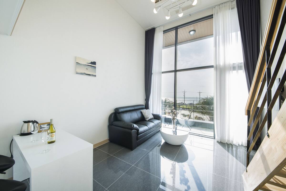 A modern living room with a black leather sofa, a white coffee table, and a large window with curtains.