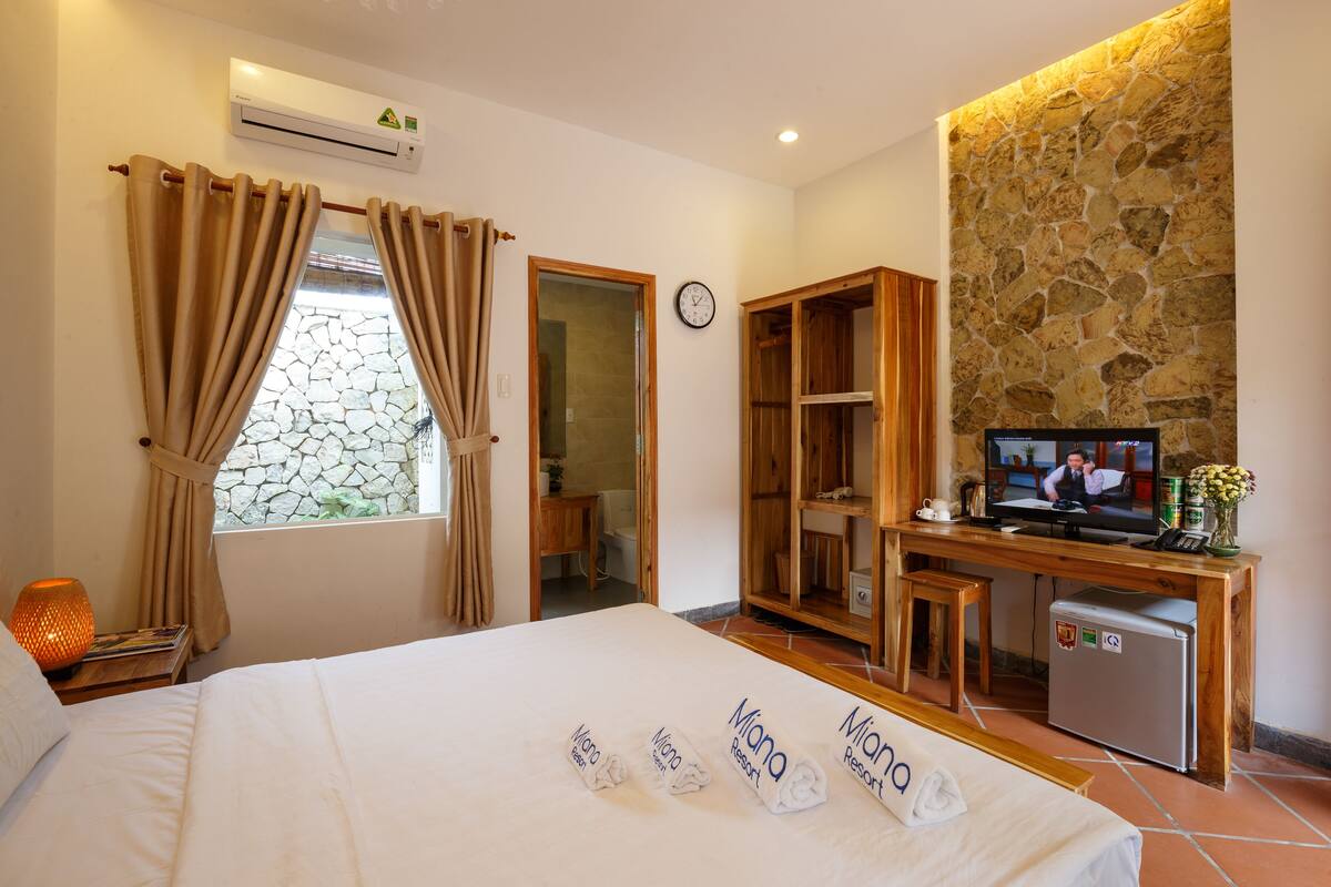 A hotel room with a bed, a desk with a TV, a small fridge, and a stone wall.
