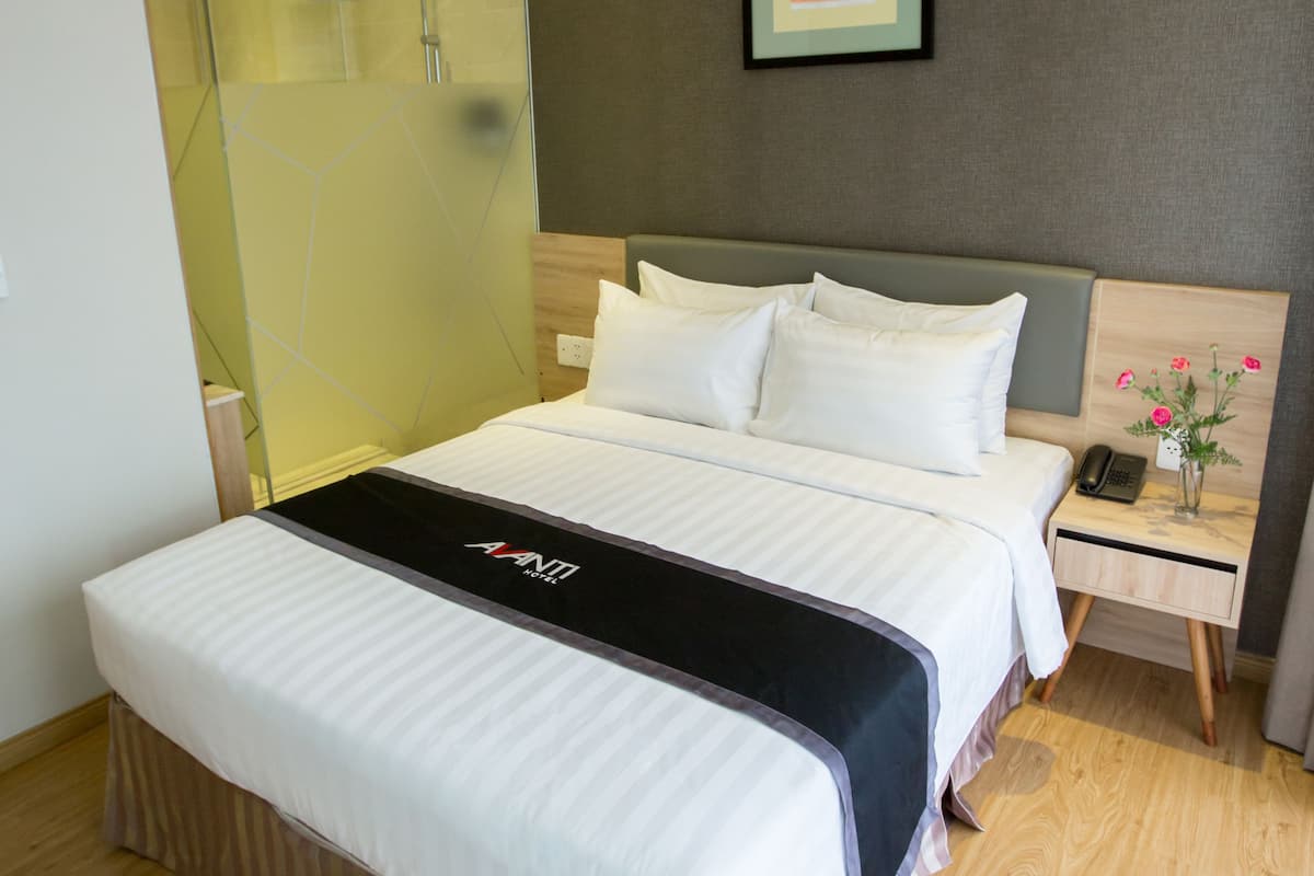 Ein modernes Hotelzimmer mit einem großen Bett, einem Nachttisch mit Telefon und Blumenvase sowie einer Duschkabine aus Glas.