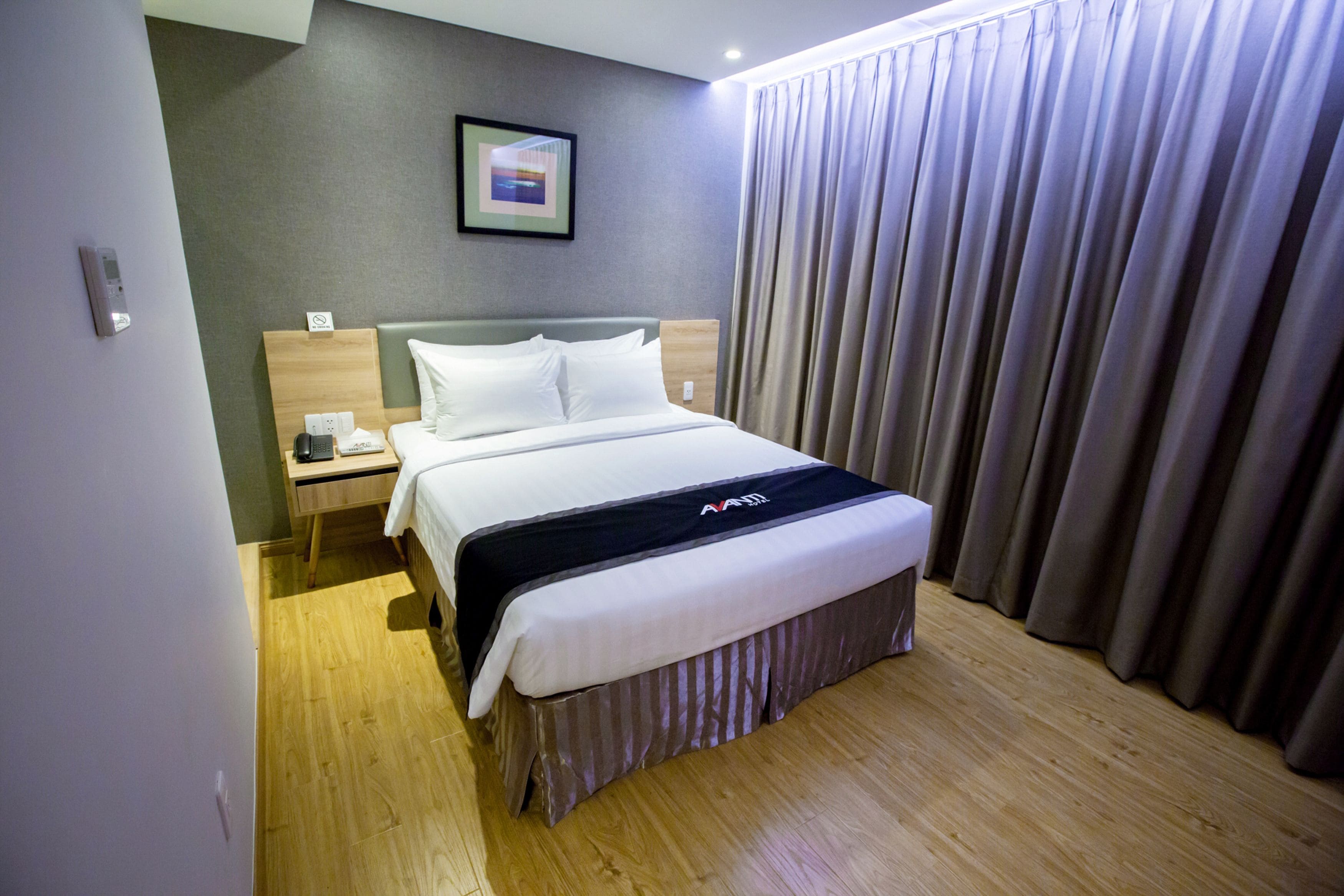 Hotellihuone, jossa on sänky, yöpöytä, seinään kiinnitetty puhelin ja ikkuna, jossa on verhot.
