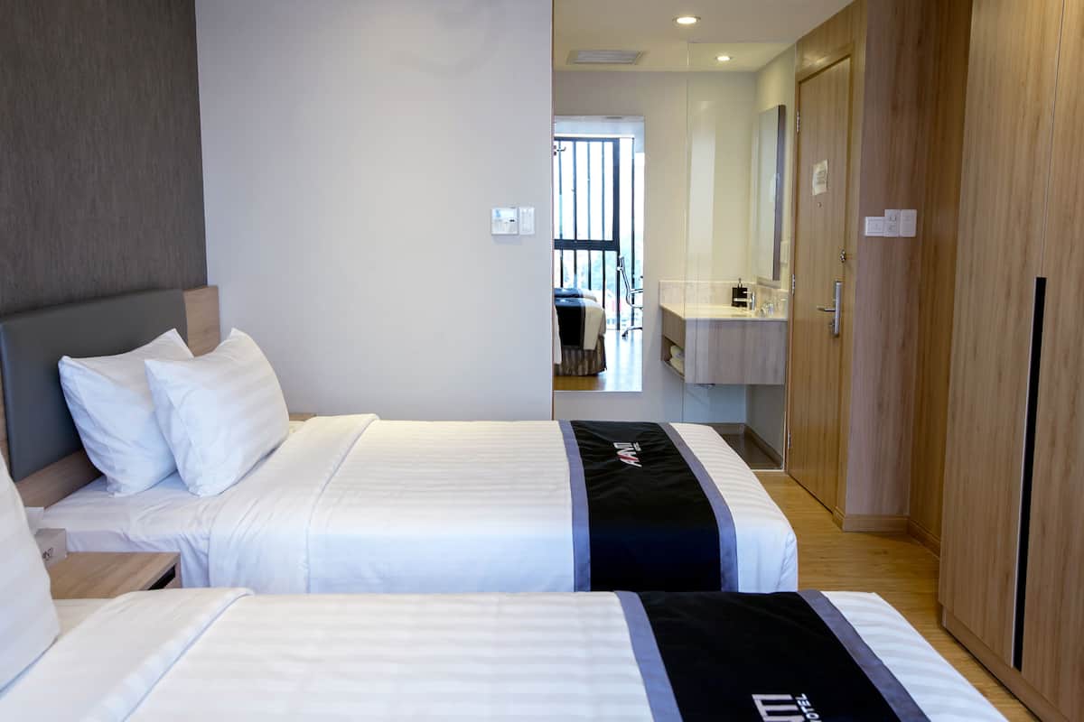 Ein Hotelzimmer mit zwei Betten, einem Holzschrank, einem Badezimmer mit Waschbecken und Spiegel sowie Blick auf den Flur.