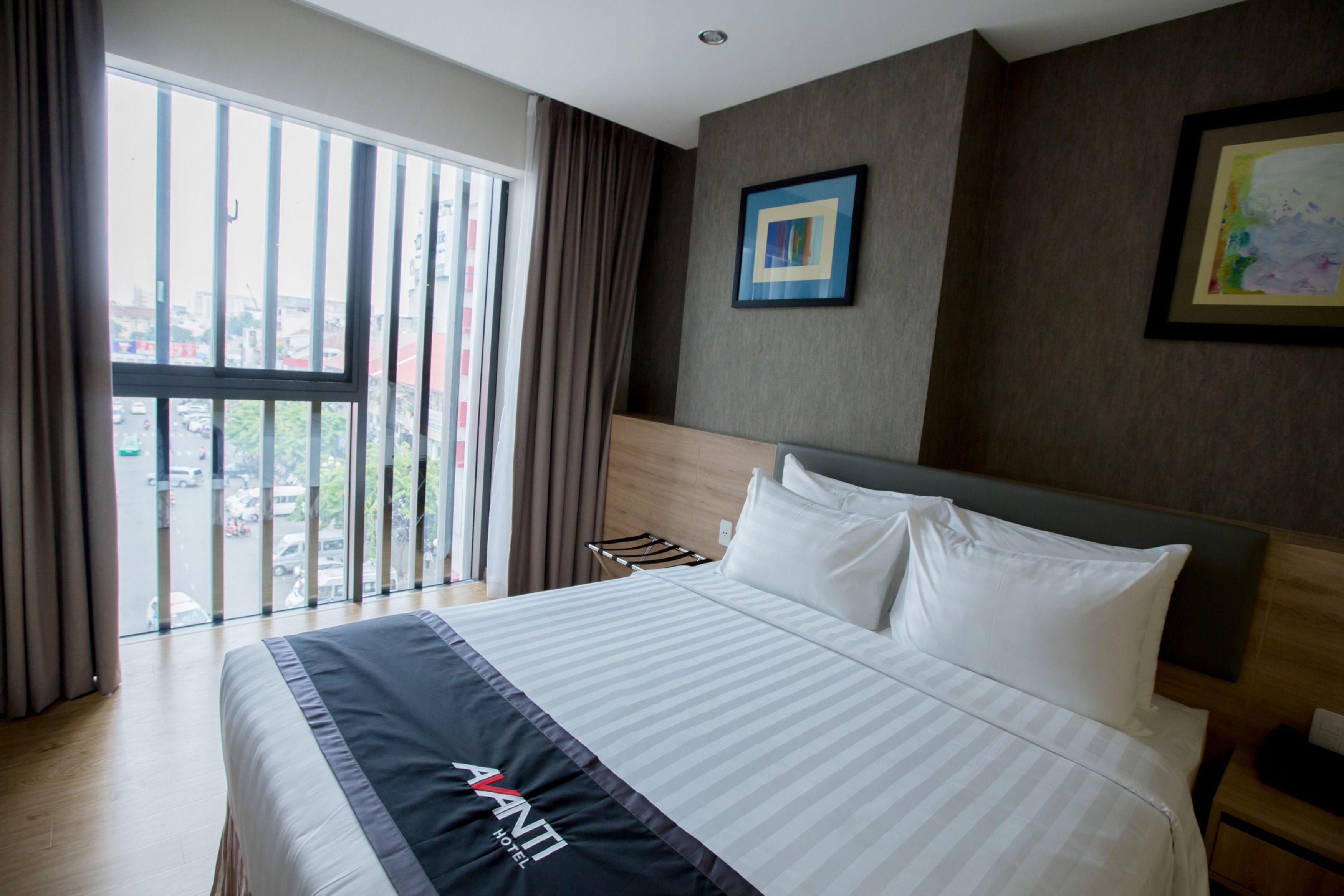 A hotel room with a large bed, a view through the window, a framed picture on the wall, and a nightstand with a lamp.