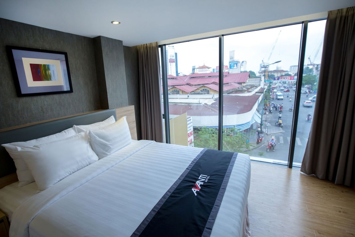 Habitación de hotel con una cama grande, un cuadro en la pared y vista a una calle concurrida.