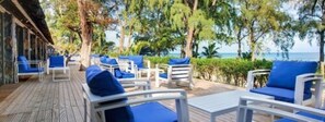 A deck with white lounge chairs and blue cushions, overlooking a beach with trees and the ocean.