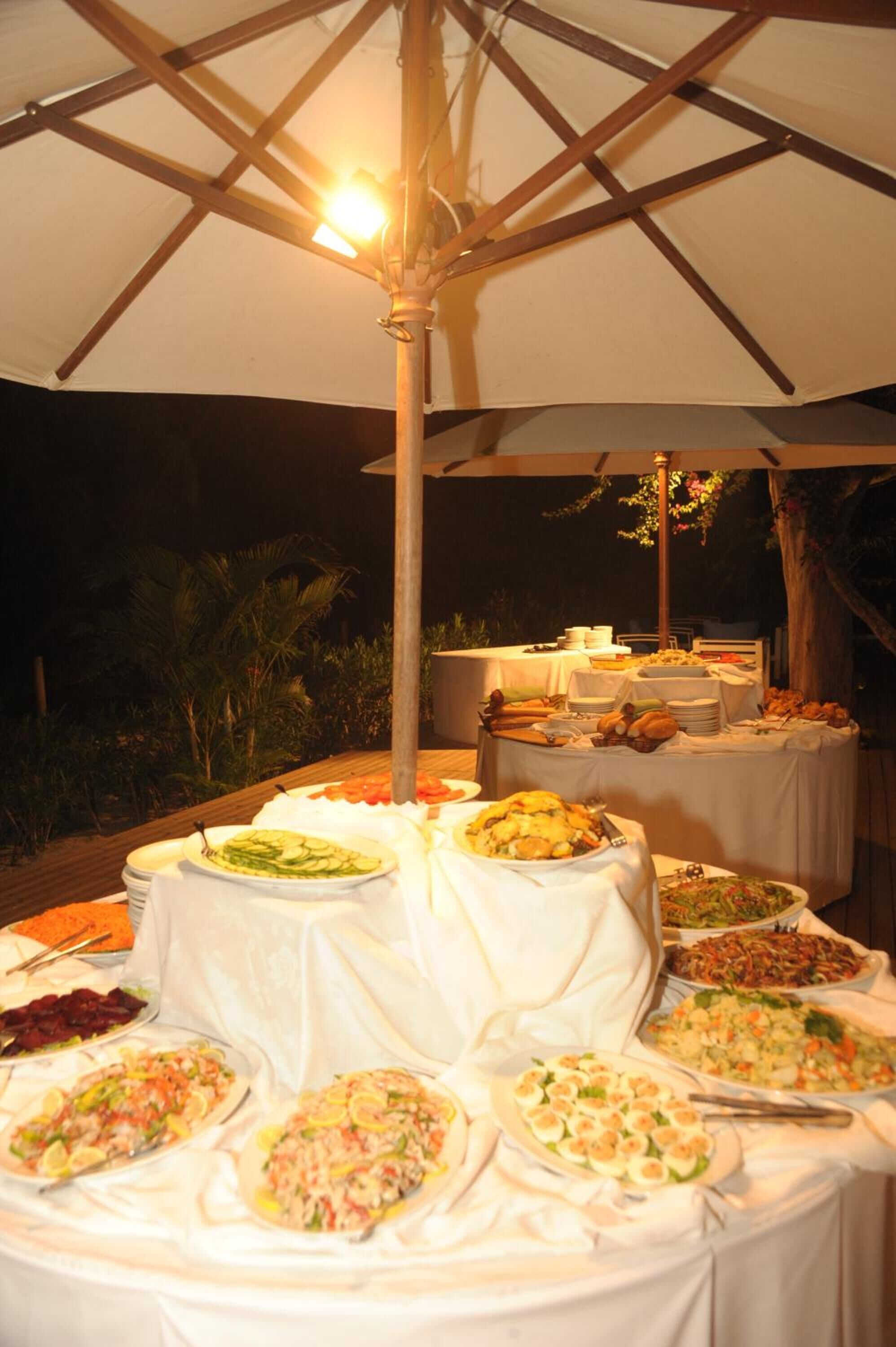 An outdoor buffet setup with multiple tables covered in white tablecloths, under a large umbrella.
