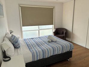 A bedroom with a bed, a chair, a bedside table, and a window with blinds. - Kangaroo Island Bayview Villas (Kingscote)