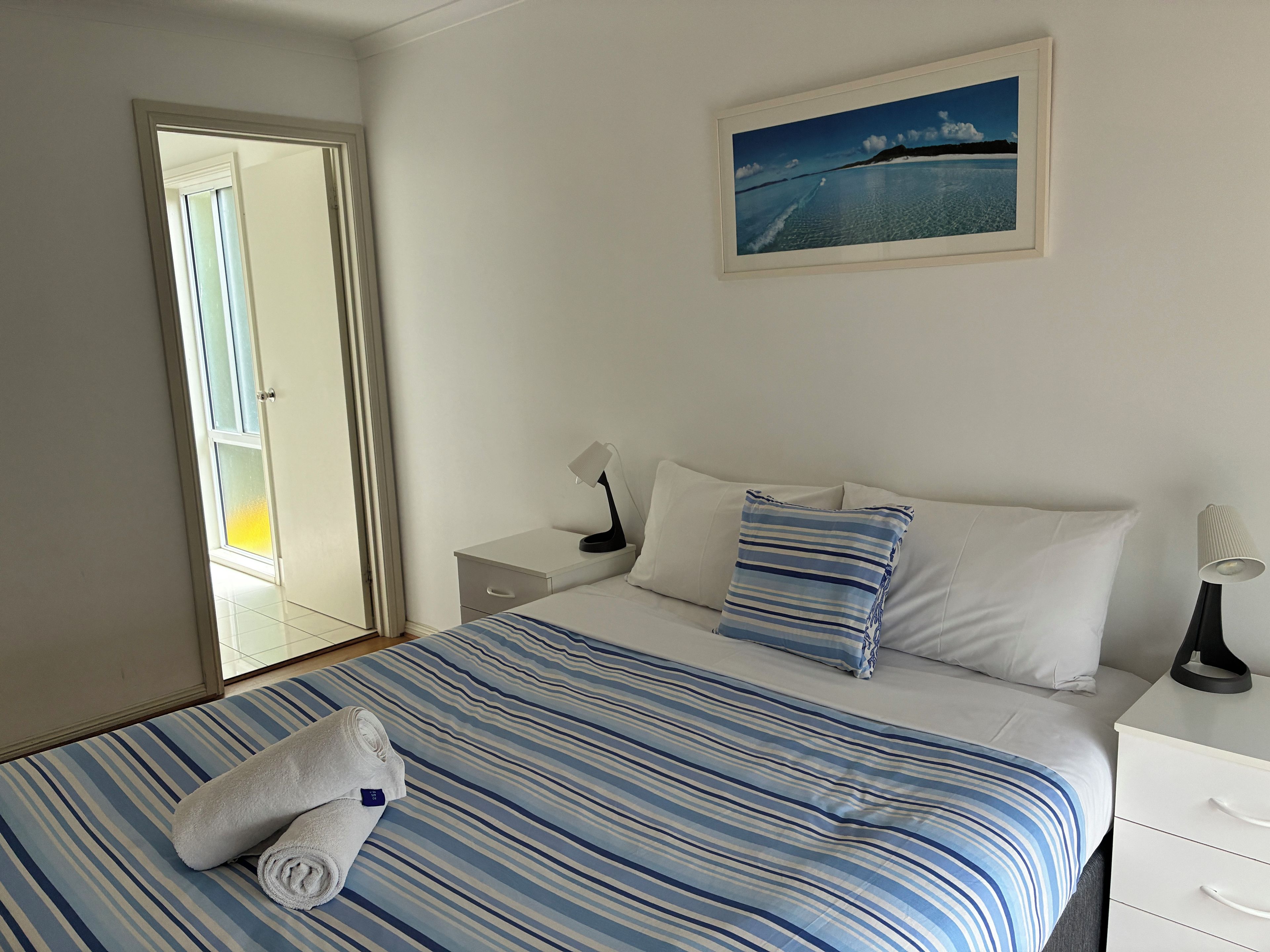 A hotel room with a bed, a nightstand, a lamp, a painting, and a view of the sea through the window.