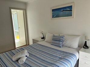 Un cuarto de hotel con una cama, una mesita de noche, una lámpara, un cuadro y vista al mar por la ventana.