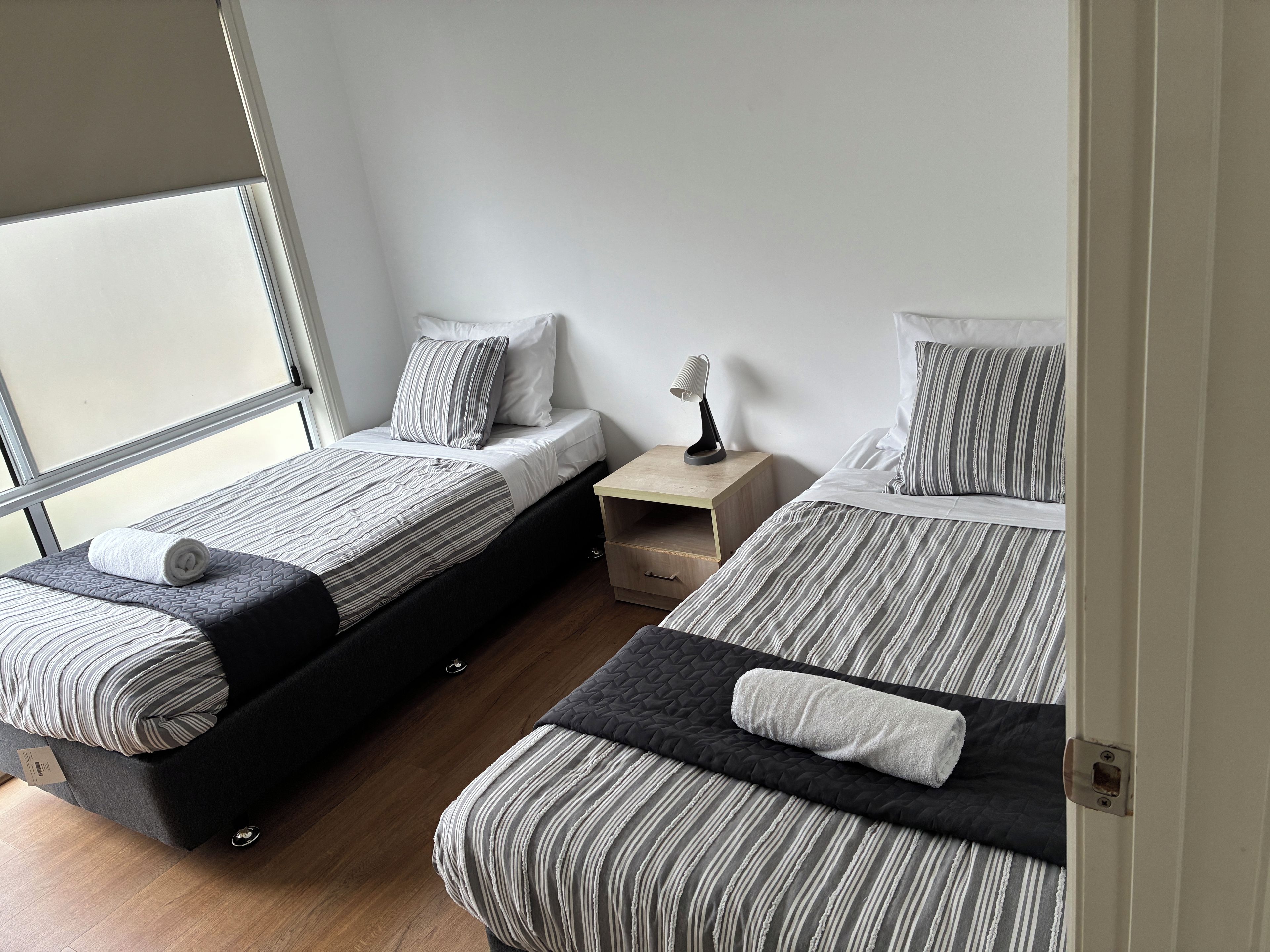 A room with two beds, each with striped bedding and pillows, a bedside table with a lamp, and a window providing natural light.