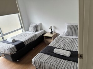 A room with two beds, each with striped bedding and pillows, a bedside table with a lamp, and a window providing natural light. - Kangaroo Island Bayview Villas (Kingscote)