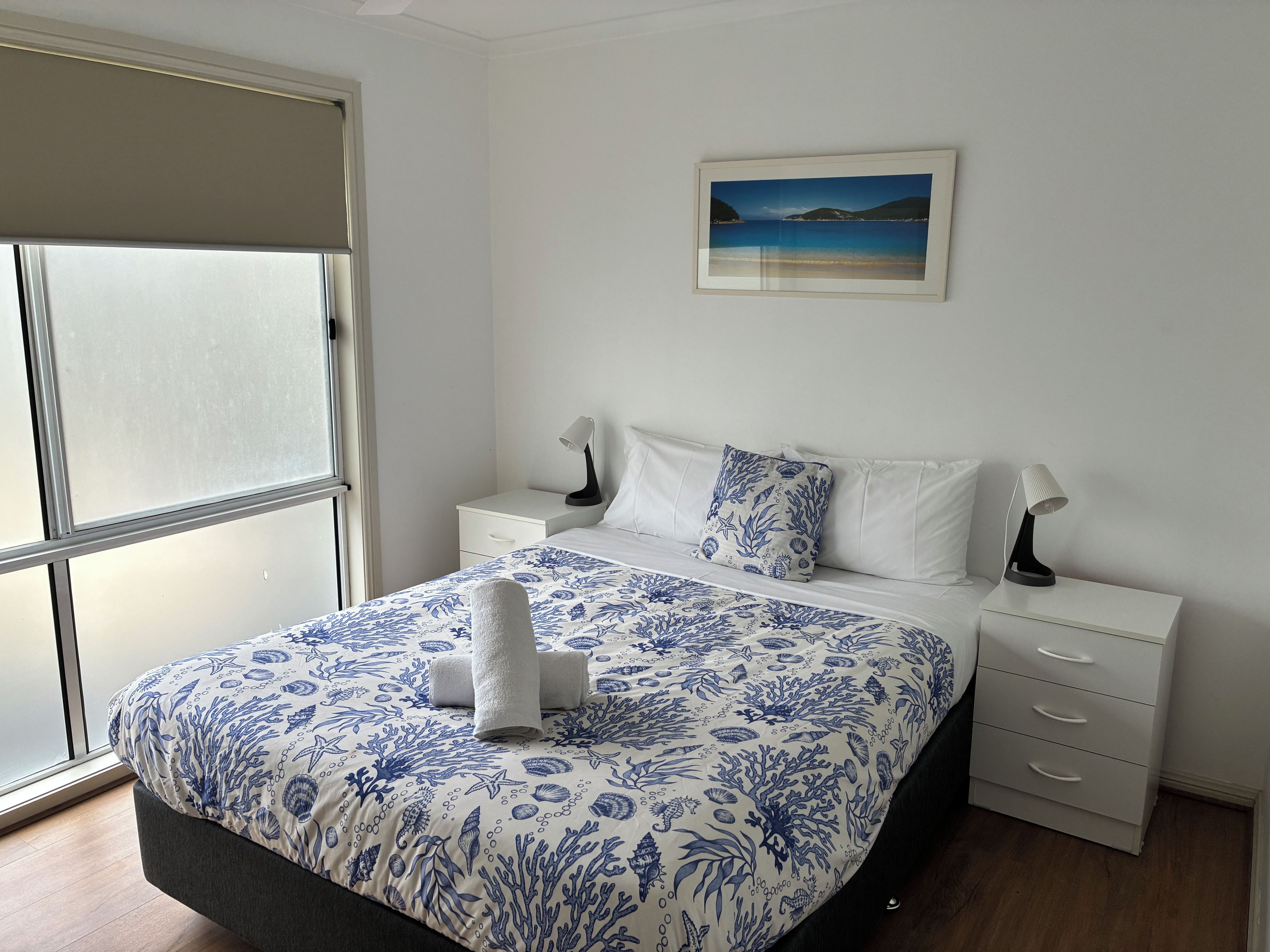 A bedroom with a bed, a nightstand, a drawer, a window, and a wall-mounted picture.