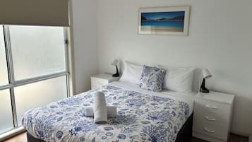 A bedroom with a bed, a nightstand, a drawer, a window, and a wall-mounted picture.