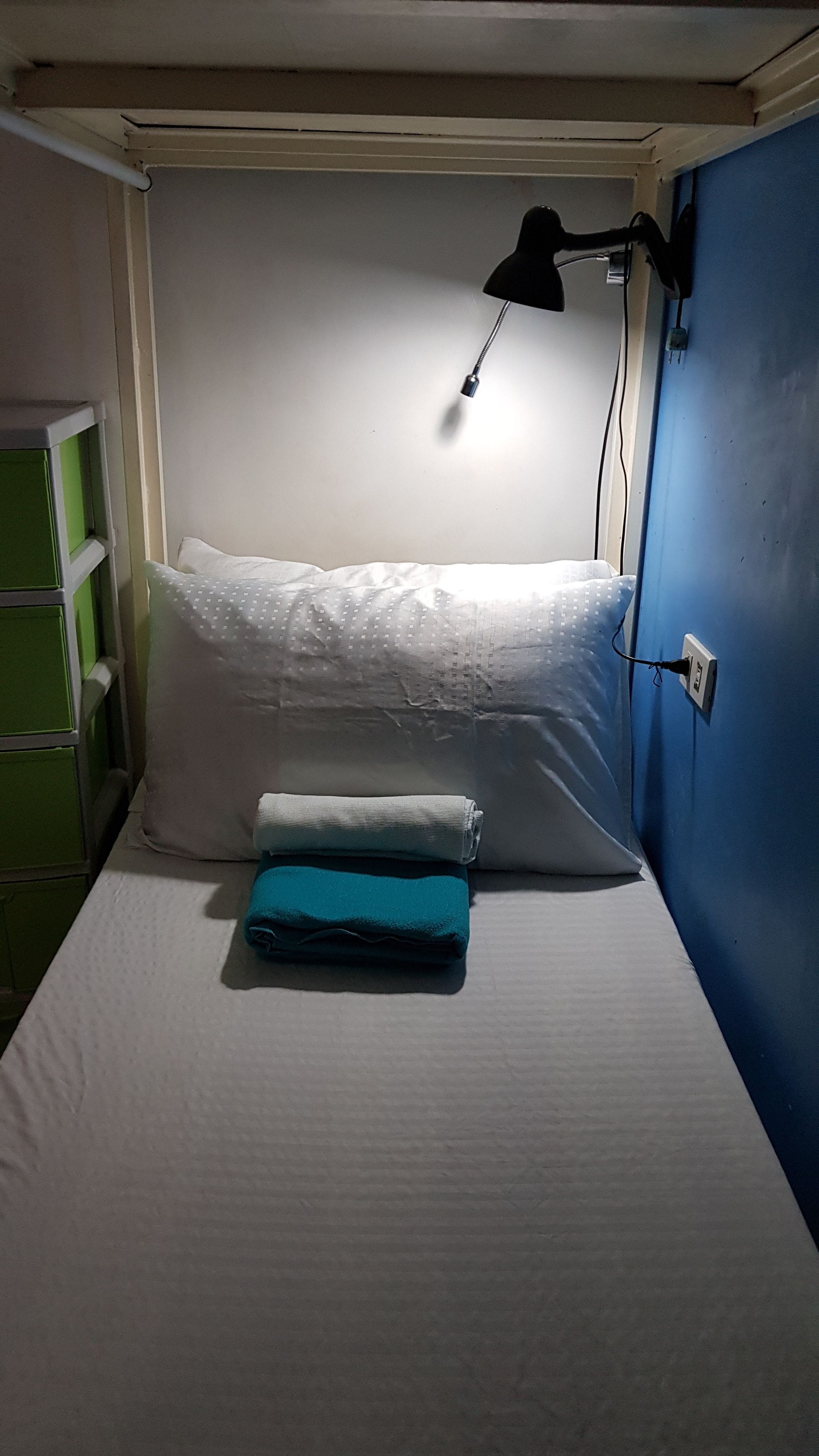 En våningssäng med en vit madrass och en turkosfärgad filt som ligger hopvikt ovanpå. En svart skrivbordslampa är monterad på väggen ovanför sängen.