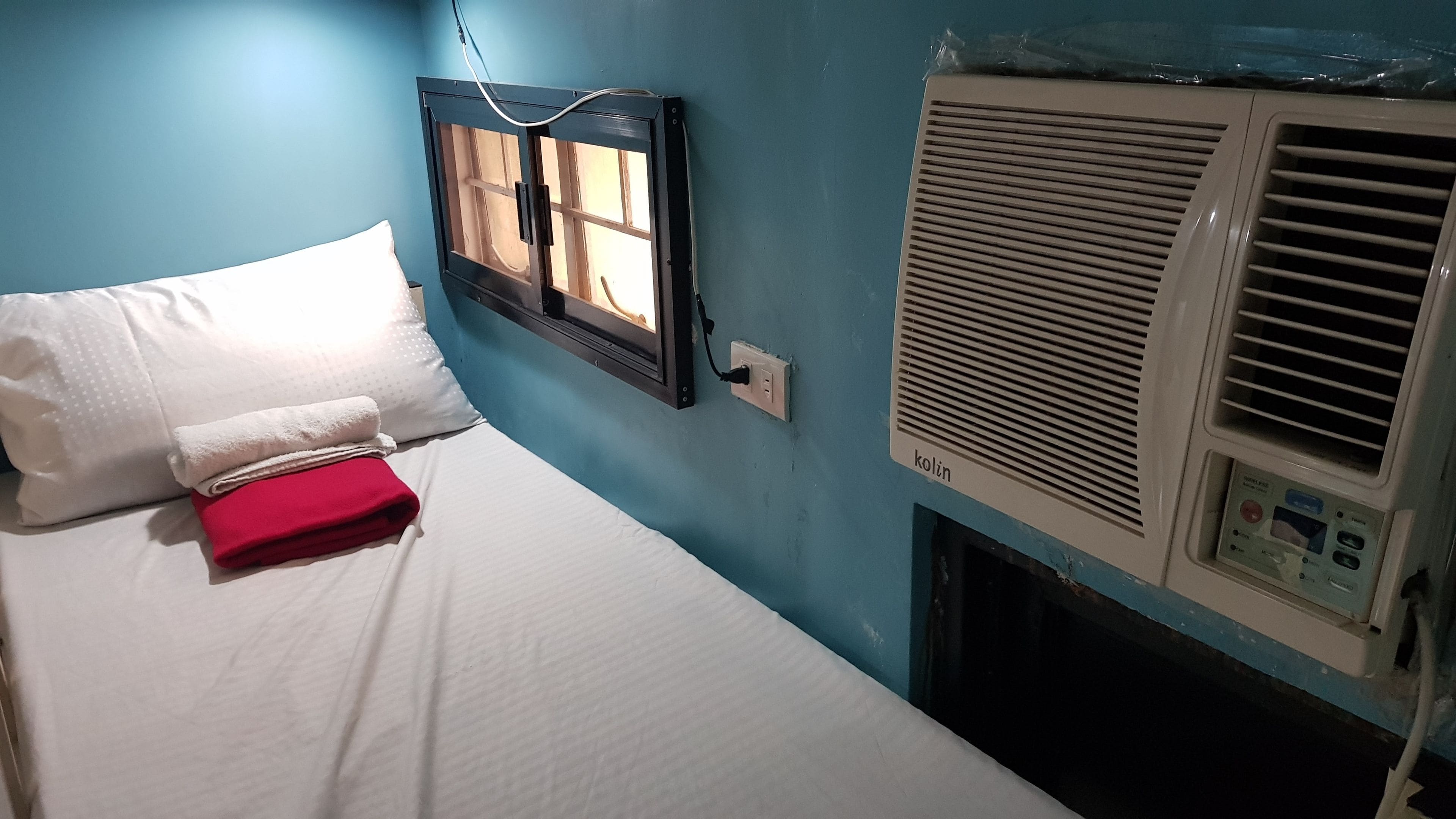 A bed with white bedding and a red blanket, a window with blinds, and an air conditioning unit on the wall.