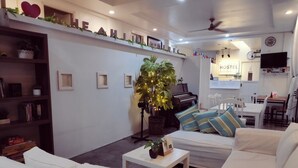 A cozy living room with white sofas, a piano, a bookshelf, and a wall-mounted sign. - Crossroads Hostel Manila (Mandaluyong)