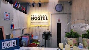 A hostel reception area with a 'CROSSROADS HOSTEL MANILA' sign, a 'WELCOME' sign, a 'FIRST AID' sign, and a 'NO ENTRY' sign. - Crossroads Hostel Manila (Mandaluyong)