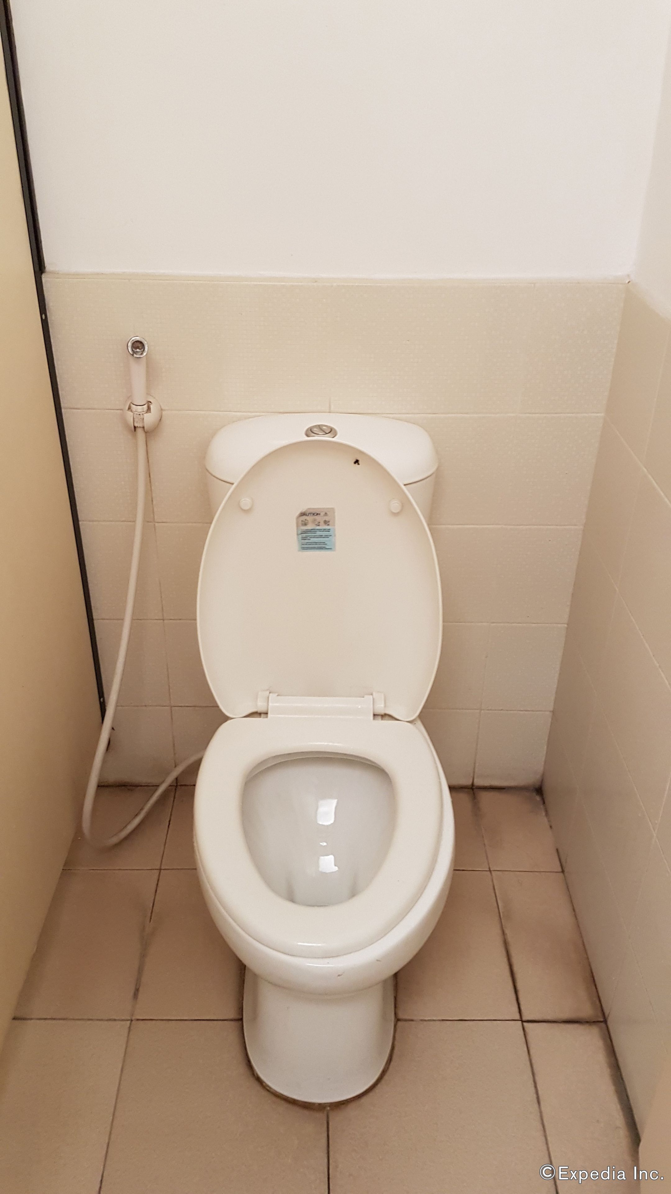 A white toilet with a flush handle and a flush button, located in a small bathroom with beige tiled walls and floor.