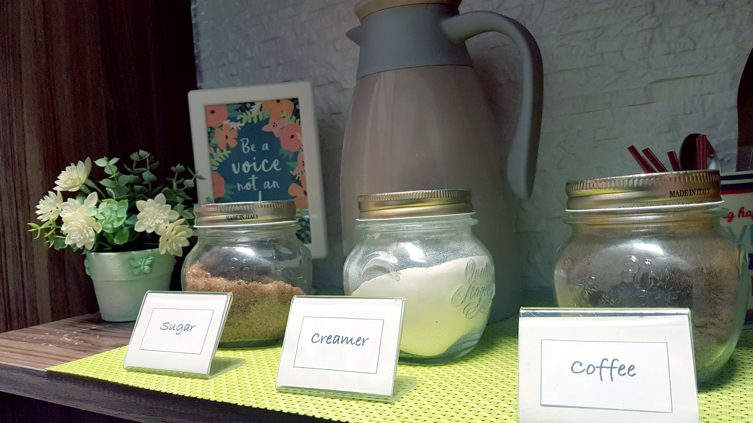 A shelf with labeled jars of sugar, cream, and coffee, a pitcher, and a sign with a quote.