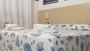 A hotel room with a bed covered in a blue and white seashell patterned bedspread, a neatly folded stack of white towels, and two framed artworks on the wall. - Il BoMa Affittacamere (Riomaggiore)