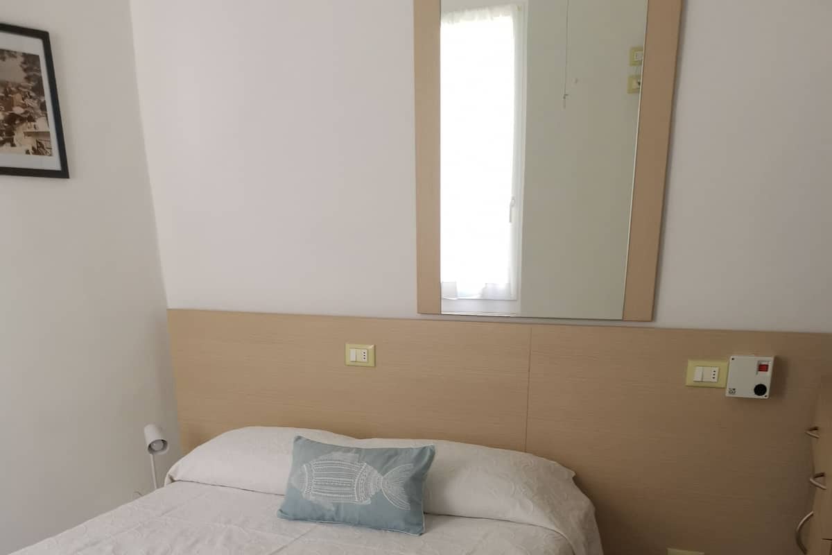 A neatly made bed with a white bedspread and a blue pillow, a bedside lamp, and a wall-mounted light switch.