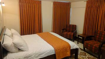 Habitación de hotel con una cama grande, dos sillas, una mesita y cortinas anaranjadas.