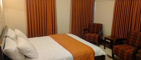 A hotel room with a large bed, two chairs, a small table, and orange curtains.