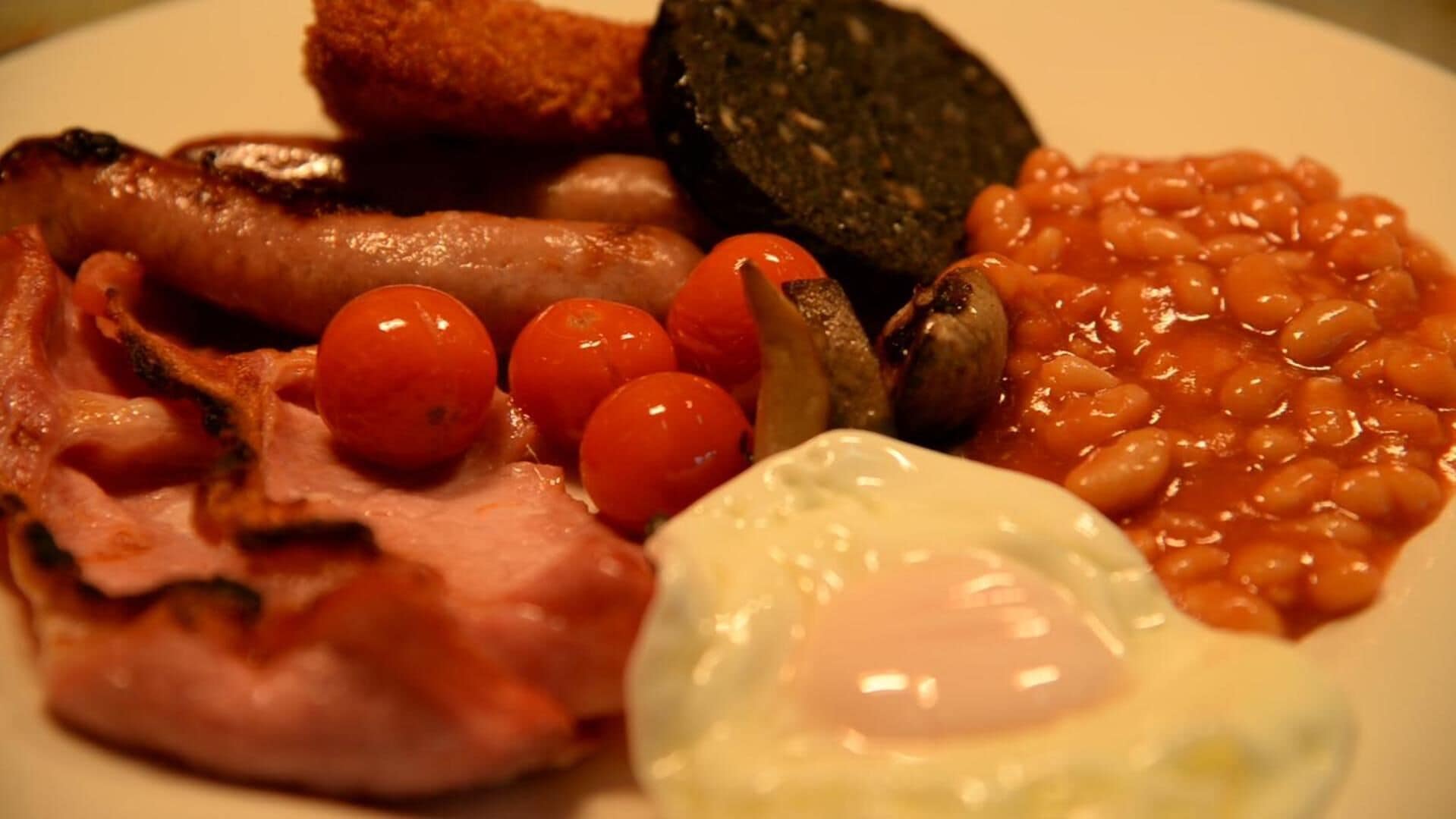 Um prato com comida de café da manhã, incluindo salsichas, ovos, tomate, feijão cozido e torradas.