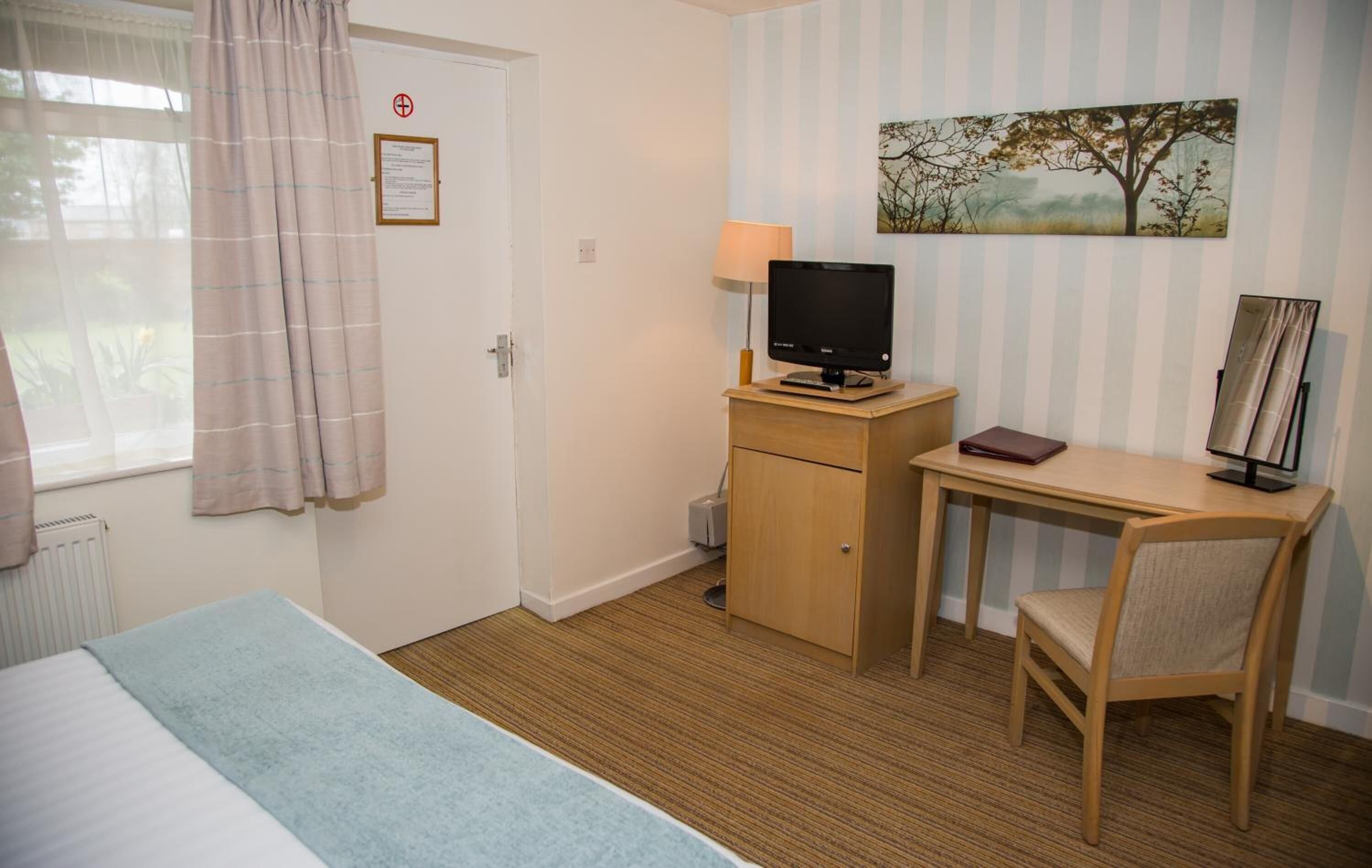 A hotel room with a bed, a desk with a computer, a chair, a window with curtains, and a painting on the wall.