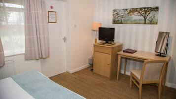 A hotel room with a bed, a desk with a computer, a chair, a window with curtains, and a painting on the wall.