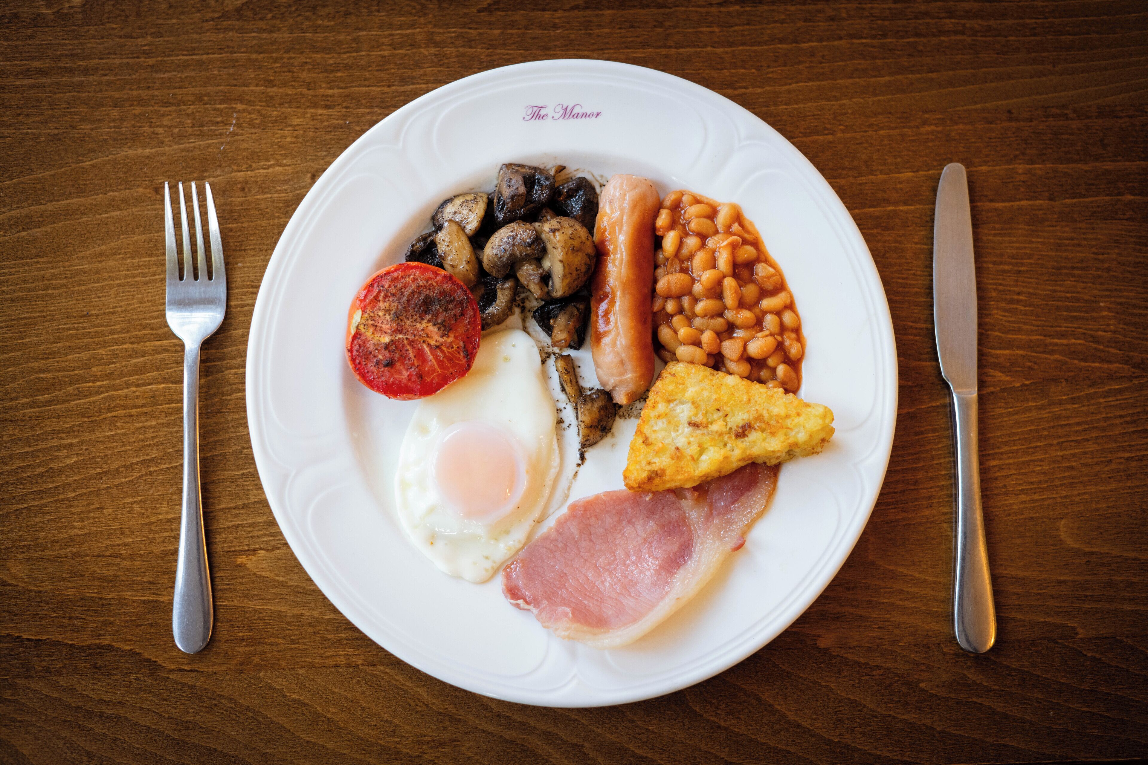 A plate of breakfast food including eggs, bacon, mushrooms, beans, and a tomato on a wooden table.