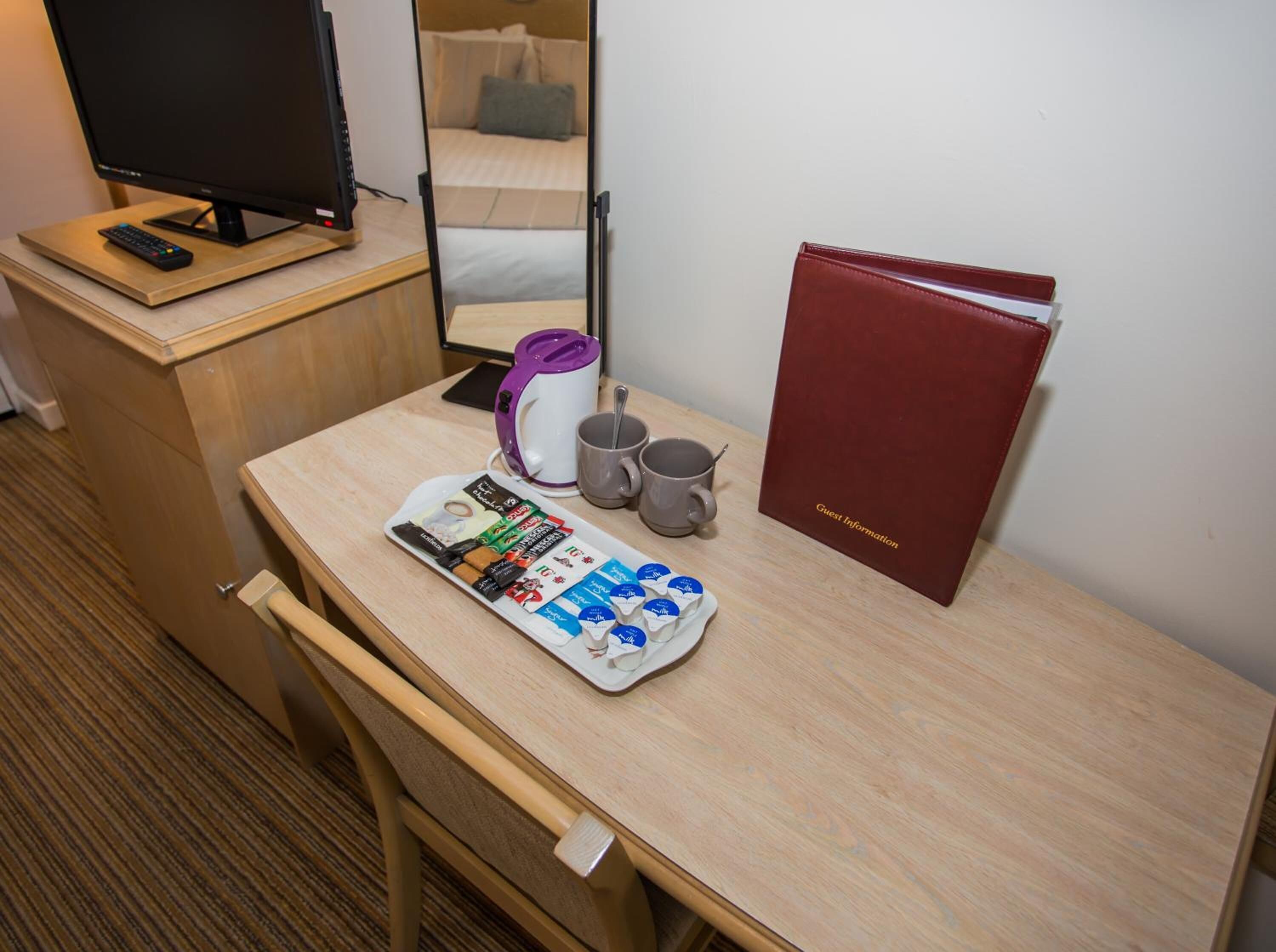 A hotel room with a wooden desk, a television, a coffee maker, a tray of coffee pods, two mugs, and a red guest information folder.