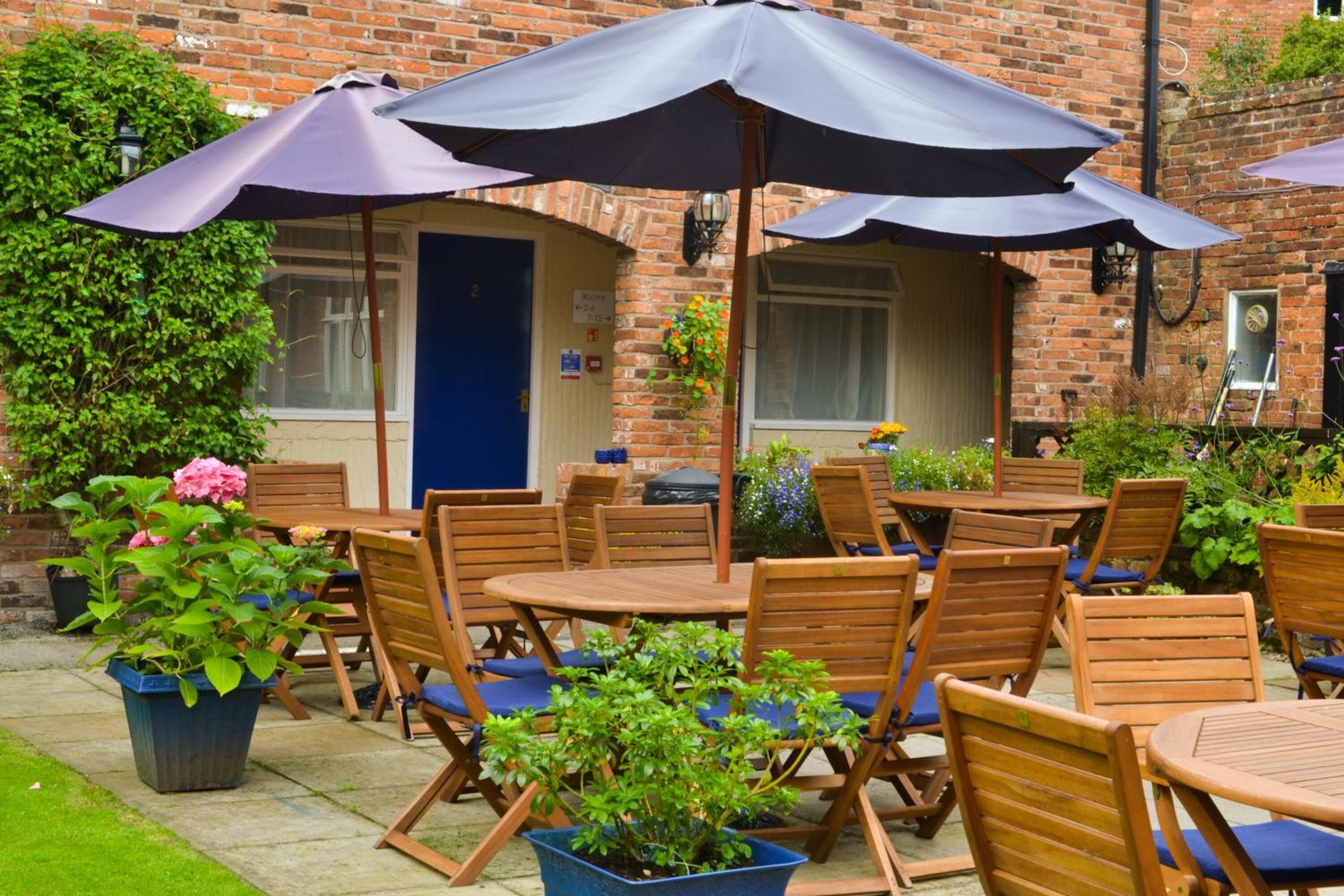 An outdoor patio with wooden tables and chairs, covered by large umbrellas, and surrounded by potted plants and a brick building.