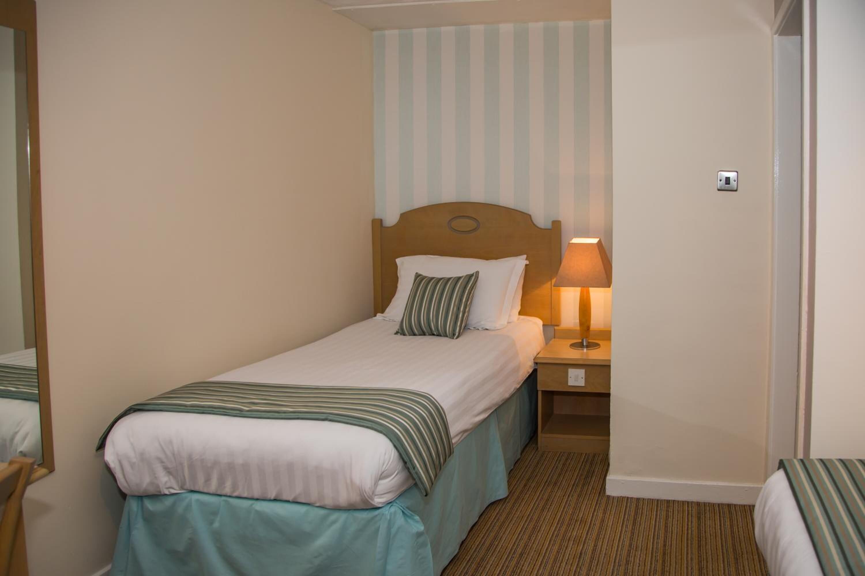 Yhden hengen sänky, jossa on puinen pääty, yöpöytälamppu ja raidallinen tyyny.