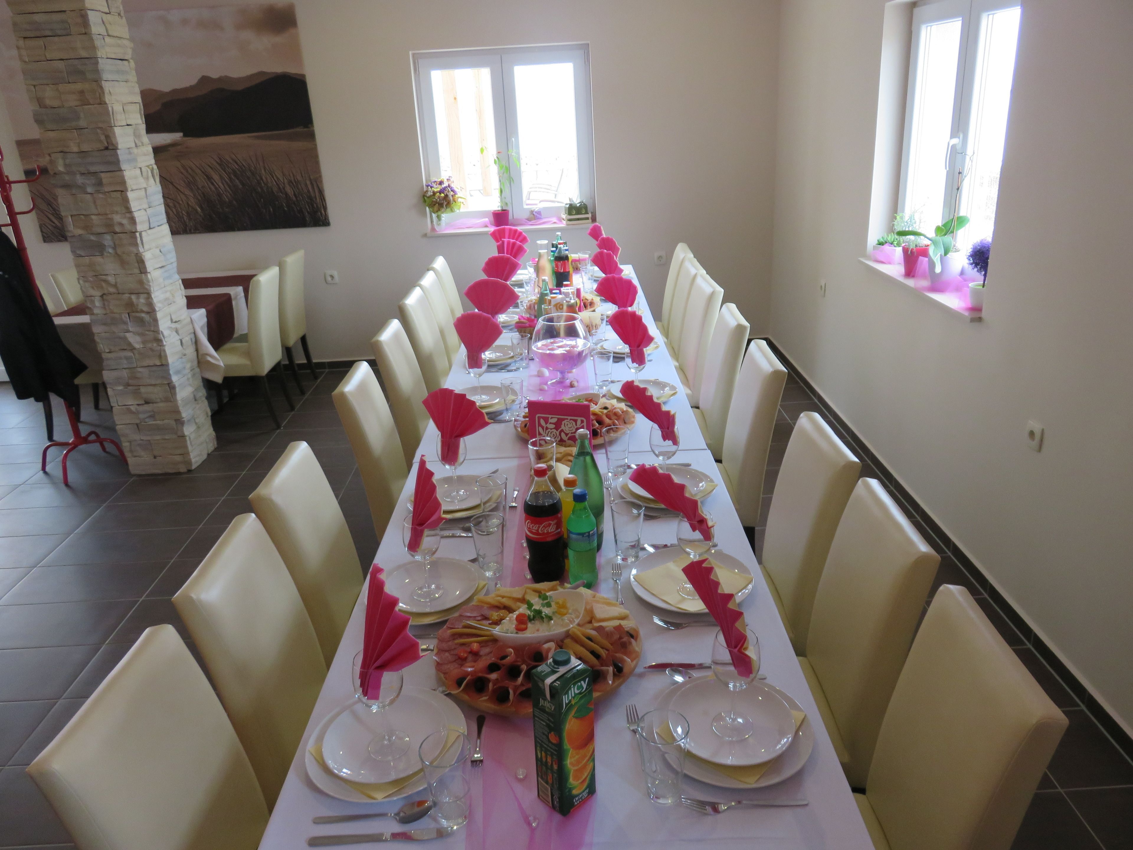 Une longue table à manger, dressée pour un repas, avec des chaises de chaque côté, un pilier en pierre et une fenêtre ornée de plantes.