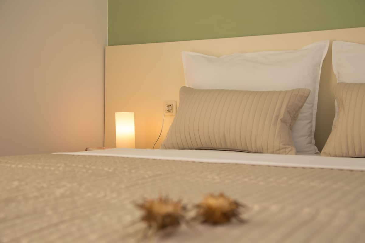 A neatly made bed with a beige bedspread and two pillows, a bedside lamp, and a power outlet on the wall.