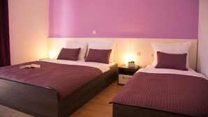 Habitación de hotel con dos camas, cada una con cubrecama morado y almohadas blancas. Hay una mesita de noche con una lámpara y una planta pequeña encima.