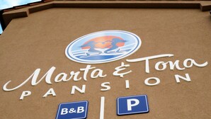 Un edificio con un letrero que dice "Marta & Tona PENSION" e incluye un logotipo con una mujer y un caballo. También hay carteles que indican "B&B" y un símbolo de estacionamiento.