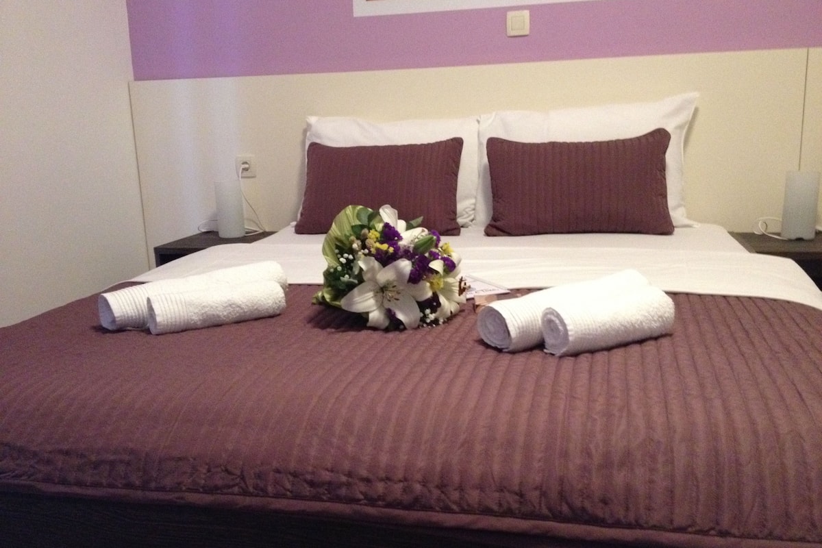 A neatly made bed with a purple bedspread, two white pillows, and a bouquet of flowers.