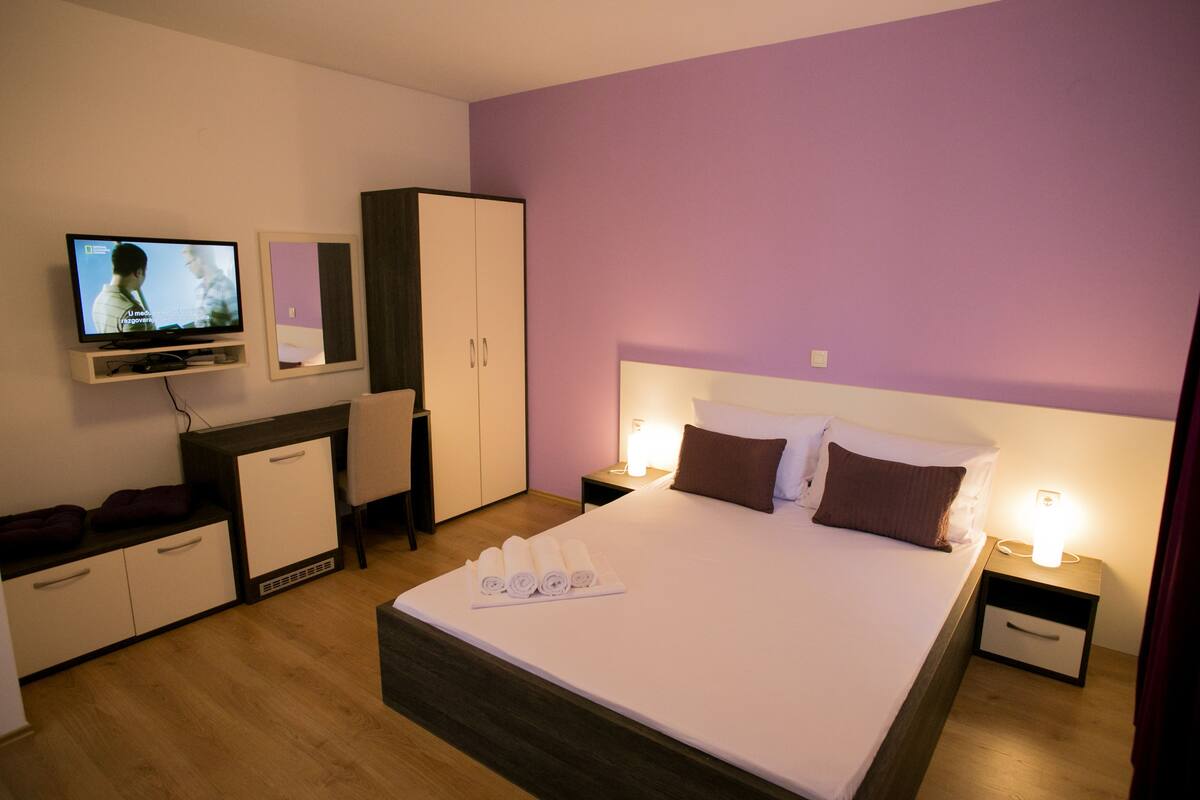 A hotel room with a bed, a TV, a wardrobe, a nightstand, and a lamp.