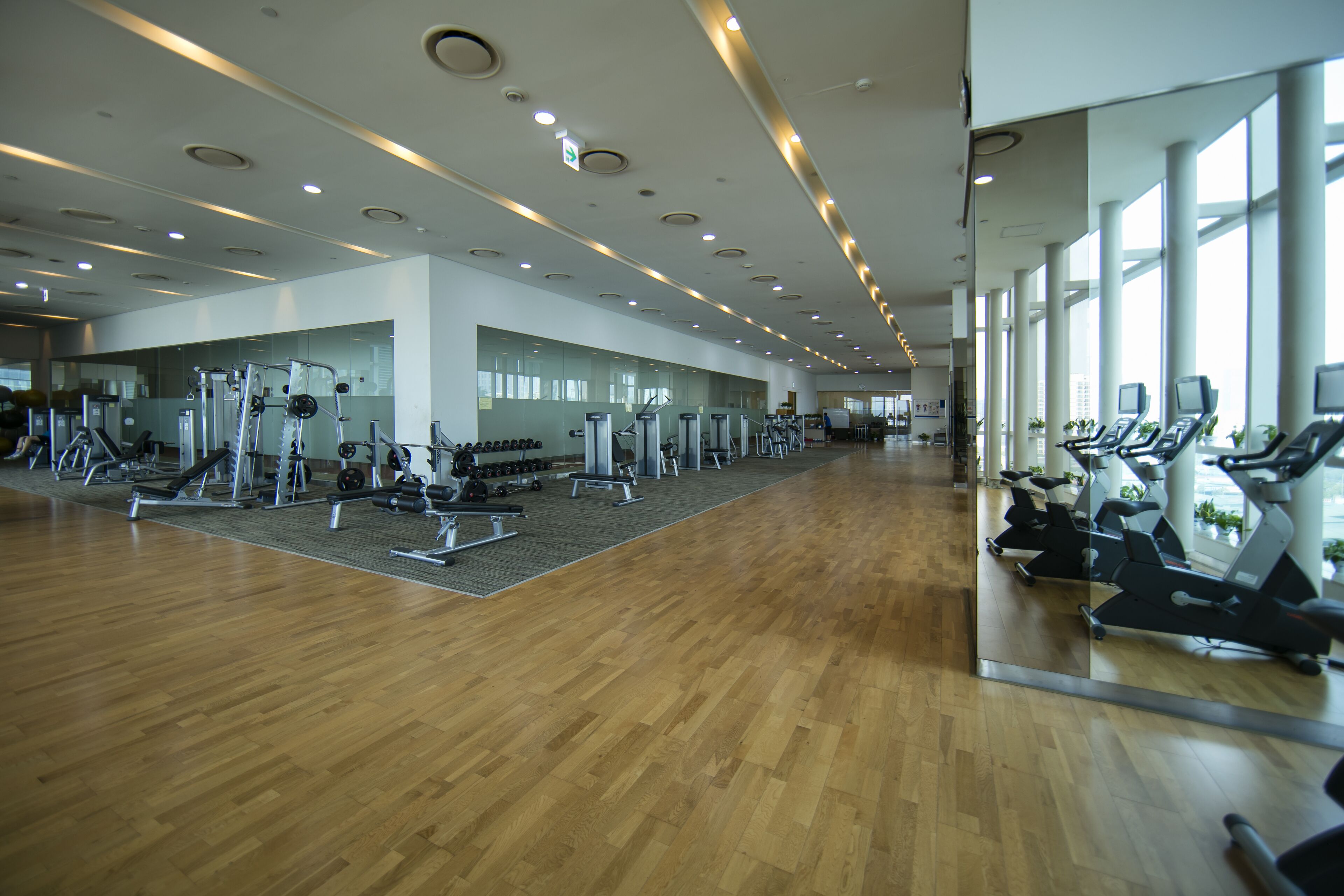 A spacious gym with various exercise equipment, large windows, and a wooden floor.