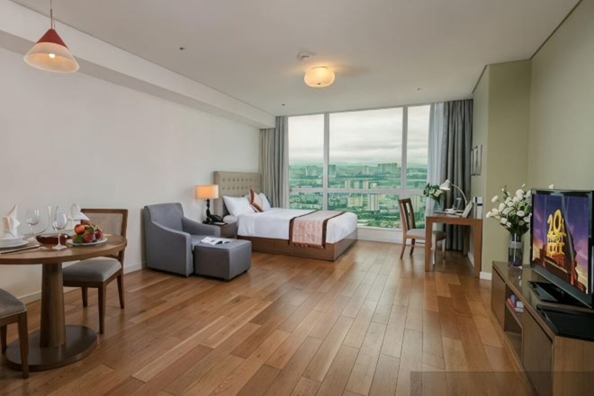 Een hotelkamer met een bed, een bureau, een stoel, een eettafel met stoelen, een televisie en een groot raam met uitzicht op de stad.